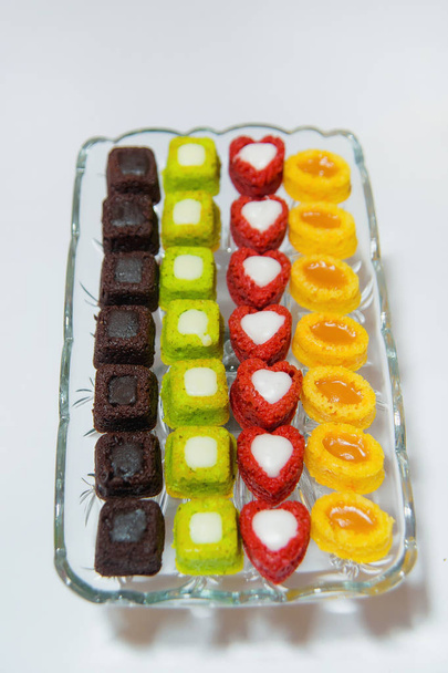 Veelkleurige mini-cakes in een vierkante glazen plaat. Taart onder de naam: Petit four. Gebak op een witte achtergrond. Zoete gebakjes. Nagerechten: Petit Fours. - Foto, afbeelding