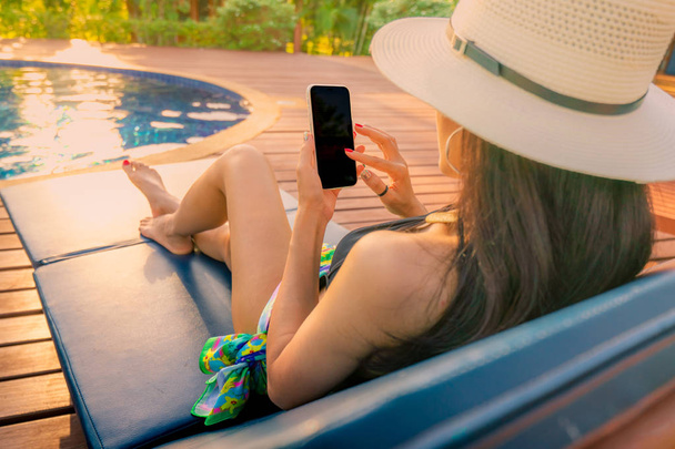 Asian woman with hat and swimsuit sitting in chair at poolside and using smartphone on summer vacation. Luxury life. Checking News on mobile phone application by swimming pool. Girl with nail manicure - Фото, зображення