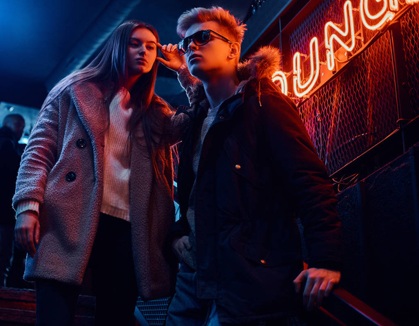 Una giovane coppia elegante indossa abiti caldi in piedi sulla scala per la discoteca sotterranea, un cartello retroilluminato sullo sfondo
 - Foto, immagini