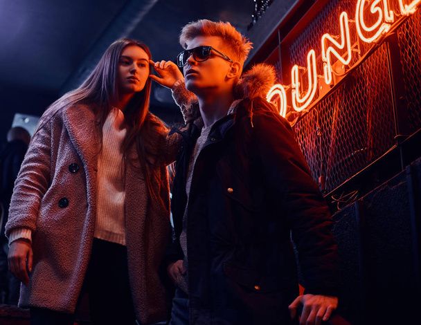 Trendy gekleed jonge paar staande op de trap naar de ondergrondse nachtclub, een verlicht bord op de achtergrond - Foto, afbeelding