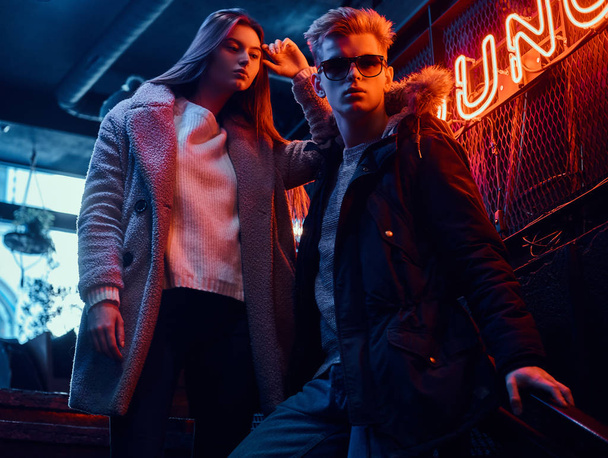 Trendy dressed young couple standing on the staircase to the underground nightclub, a backlit signboard in the background - Foto, imagen