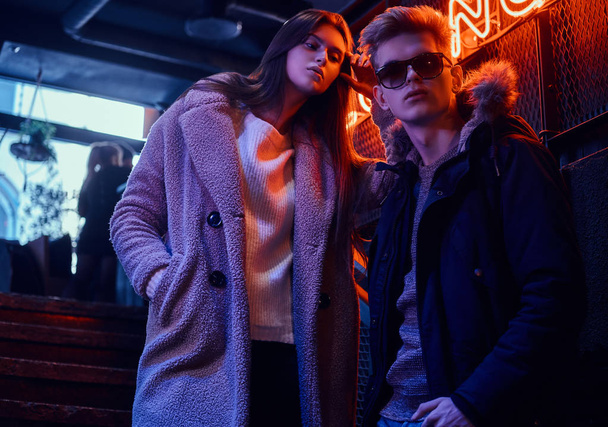 Um jovem casal elegante vestindo roupas quentes em pé na escada para a discoteca subterrânea, uma placa retroiluminada no fundo
 - Foto, Imagem
