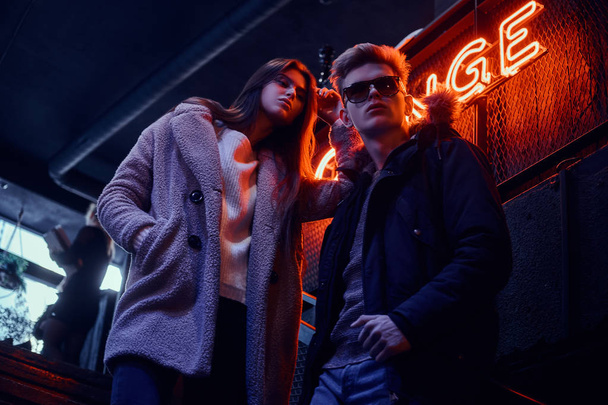Молодая стильная пара в теплой одежде, стоящая на лестнице в подземный ночной клуб, на заднем плане вывеска с подсветкой
 - Фото, изображение