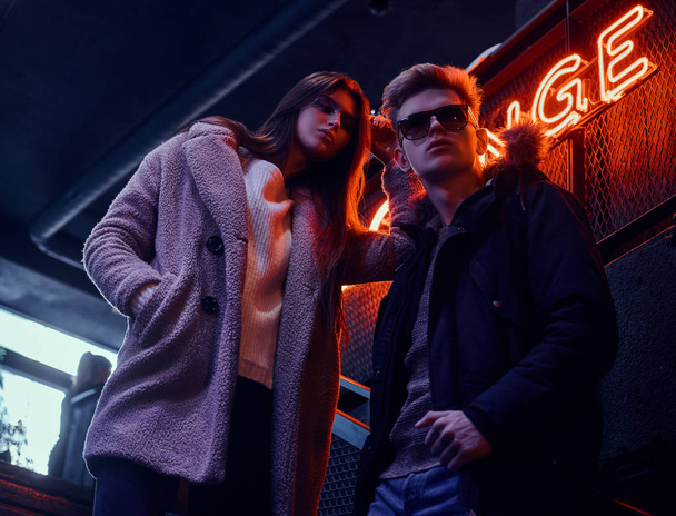 ein junges stylisches Paar in warmer Kleidung steht auf der Treppe zum Nachtclub, im Hintergrund ein hinterleuchtetes Schild - Foto, Bild