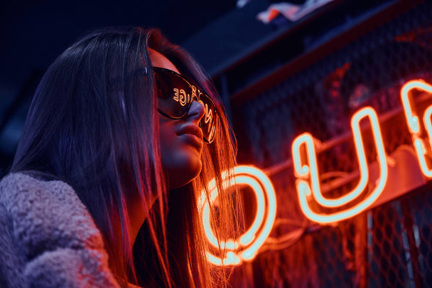 Close-up portrait of a sensual young girl wearing sunglasses and coat standing at the cafe with the industrial interior, a backlit signboard in the background - Фото, изображение