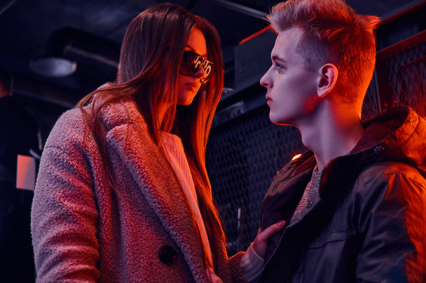 A young stylish couple passionately look at each other while standing in the cafe with industrial interior, a backlit signboard in the background - Photo, Image