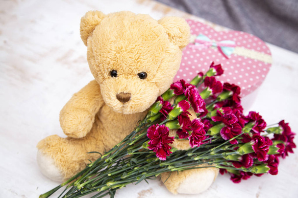 Cute toy - bear with burgundy carnations and a box in the shape of a heart, a gift, on light background  - Foto, Imagen