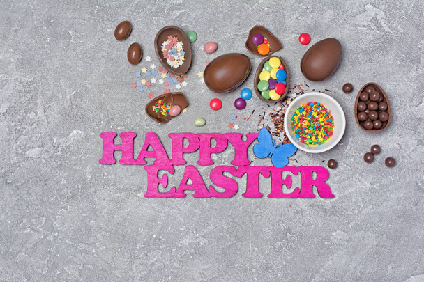 Composition of pink text happy easter with chocolate traditional eggs and bright colorful dragee with sugar sprinkles or confetti on gray concrete background - Фото, изображение