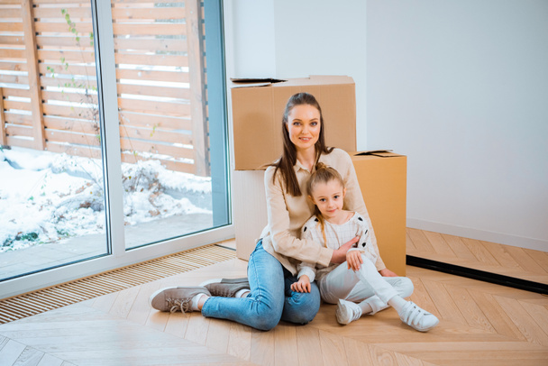 cheerful mother embracing cute daughter while sitting on floor in new home - Photo, Image