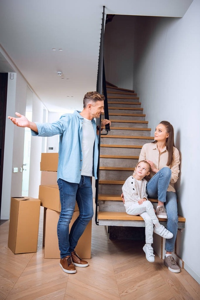 handsome man looking at cute daughter and wife sitting on stairs in new home  - Photo, Image