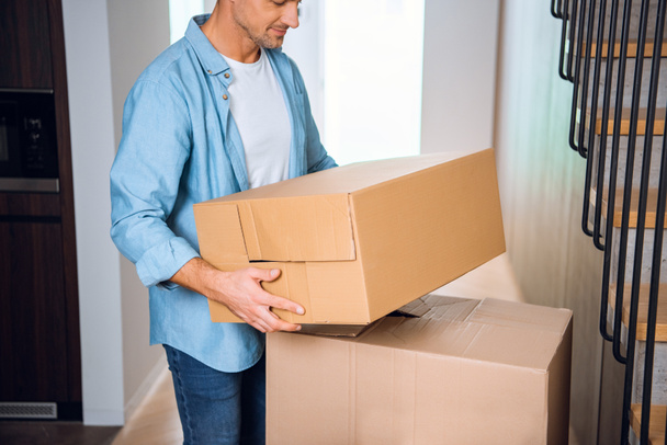 cropped view of man holding box in hands and smiling in new home  - Foto, Bild
