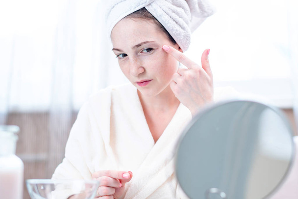Красивая счастливая молодая женщина с полотенцем на голове, смотрящая на свою кожу в зеркало. Концепция своевременной гигиены и ухода за кожей дома
 - Фото, изображение