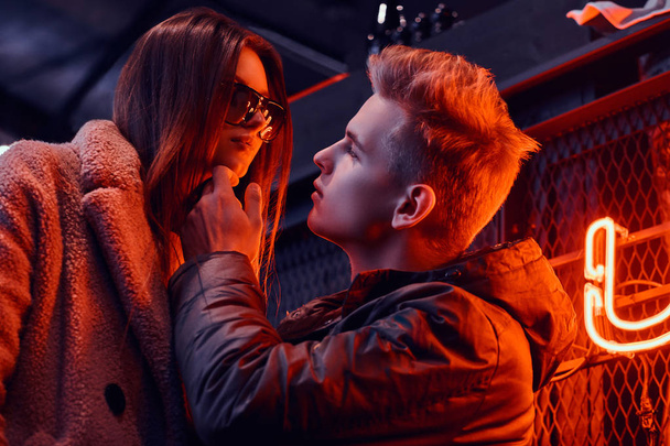 Close-up portrait of a passionate young couple looking at each other in the underground nightclub with industrial interior, he gently touches her chin - Foto, imagen