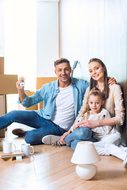 cheerful man sitting on floor with house shaped key chain near wife and daughter  - Foto, Bild