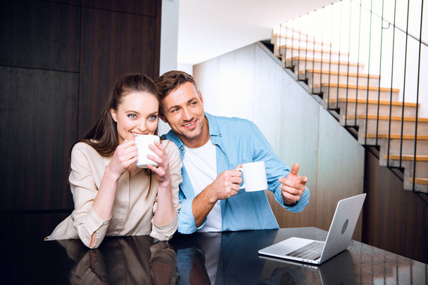 cheerful man pointing with finger at laptop near wife drinking coffee - Photo, Image