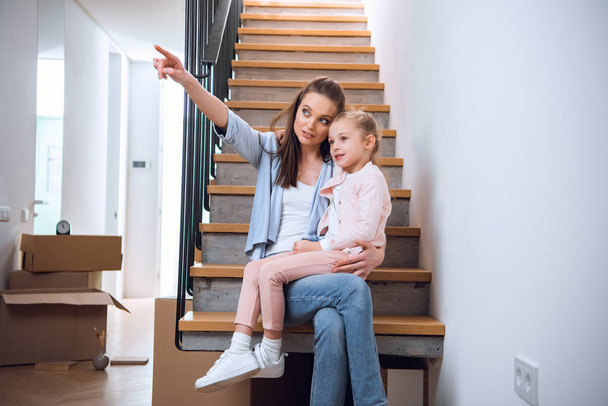 attractive woman sitting with cute daughter on stairs and pointing with finger in new home  - Photo, Image