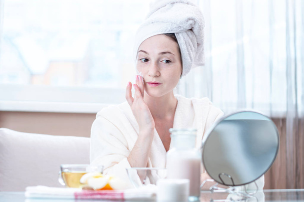 Mooie gelukkig jonge vrouw met een handdoek op haar hoofd cream voor natuurlijke cosmetica op gezicht hydraterende de huid toe te passen. Het concept van hygiëne en verzorging voor de huid thuis - Foto, afbeelding