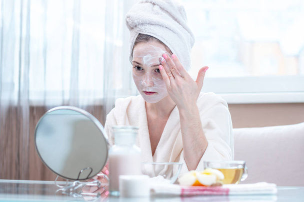 Mulher jovem feliz bonita com uma toalha na cabeça aplicando creme de cosméticos naturais no rosto hidratando a pele. O conceito de higiene e cuidado da pele em casa
 - Foto, Imagem