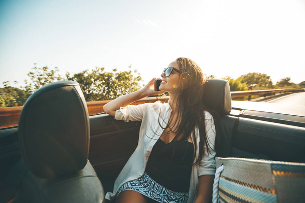Молодая женщина пользуется мобильным телефоном и сидит в кабриолете в летний день
 - Фото, изображение