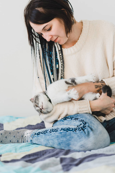 Pretty smiling young dreadlock girl with a little kitten in her hands in the cozy bedroom   - 写真・画像