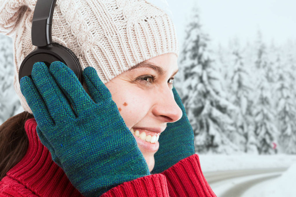 Close-up of woman touching headphones with green gloves having happy expression as enjoying music in winter snowy background - Photo, image