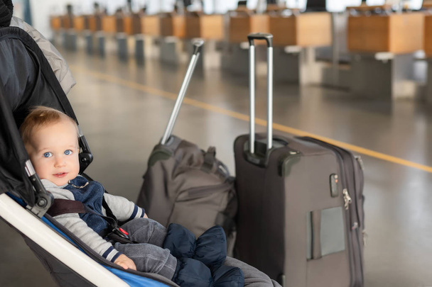 Netter lustiger kaukasischer Junge, der im Kinderwagen in der Nähe des Gepäcks am Flughafenterminal sitzt. Kinderwagen mit Koffern in der Nähe des Check-in Schalters. Reisen mit kleinen Kindern. - Foto, Bild