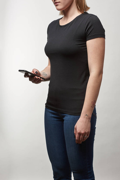 częściowy widok młodej kobiety w czarny t-shirt z kopii miejsca za pomocą smartfona na białym tle - Zdjęcie, obraz