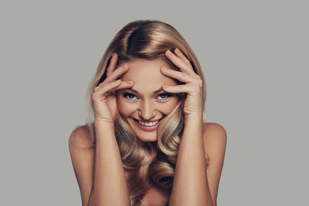blonde young woman touching face with hands and smiling while standing against grey background - Photo, image