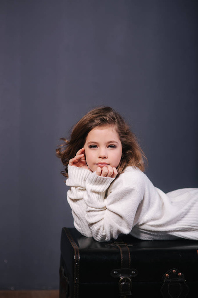 kleines schönes Mädchen mit braunen Haaren in weißem Pullover. Studio. eine Prinzessin posiert für einen Fotografen. Make-up und Frisur - Foto, Bild