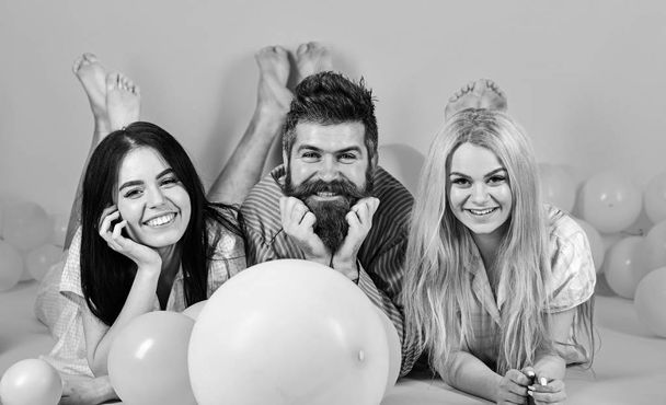 Blonde and brunette on smiling faces have fun with bearded macho. Best friends, lovers near balloons, pink background. Lovers or best friends in pajamas at girlish bedroom party. Gossip concept - Foto, Imagen