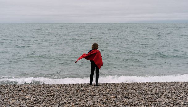 Rückansicht einer Reisenden mit rotem Umhang, die am Meeresstrand steht, träumt und schöne Landschaften genießt. Reisekonzept. Urlaub.  - Foto, Bild