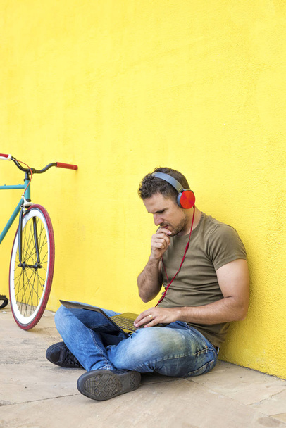 Vue de face d'un gars attrayant est assis sur le sol avec un regard à la mode, écrire dans un ordinateur portable en utilisant un ordinateur, avoir des écouteurs tout en regardant l'écran contre un mur jaune
 - Photo, image