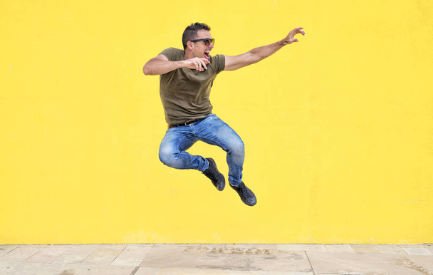 Front view of a young happy man wearing sunglasses jumping against a yellow bright wall in a sunny day - Foto, Imagem