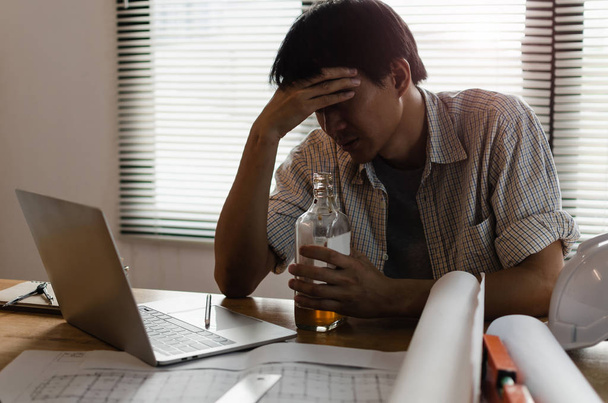young asian engineer, architect, contractor thinking and worrying about new project contract plan with laptop and bottle of alcohol on desk in office center at construction site, alcoholism concept - Photo, image