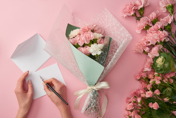 abgeschnittene Aufnahme einer Frau, die auf eine Grußkarte schreibt und einen schönen rosafarbenen Blumenstrauß isoliert auf rosa - Foto, Bild