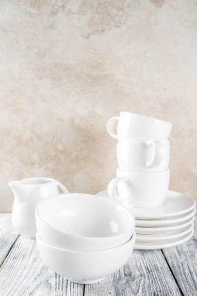 Ποικιλία στοίβα καθαρό άδειο νέο λευκό μαγειρικά σκεύη, πλάκες, μπολ, κούπες κύπελλα. Σε λευκό φόντο σκυροδέματος - Φωτογραφία, εικόνα