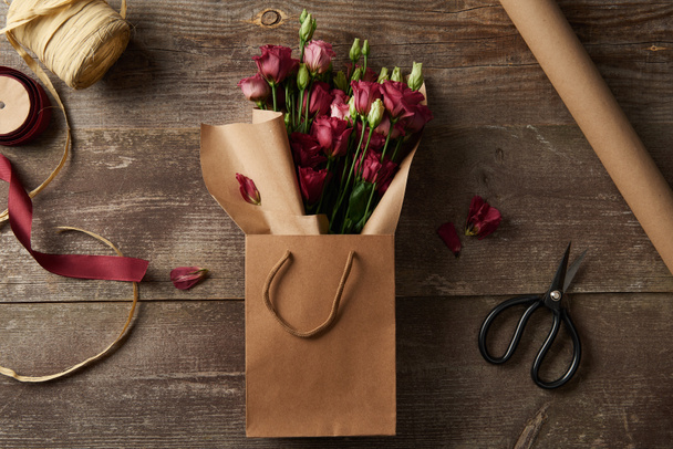 vista superior de belas flores de eustoma vermelho em saco de papel, tesoura, papel artesanal e fitas na superfície de madeira
 - Foto, Imagem