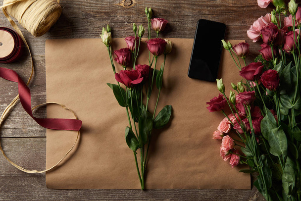 το top view από όμορφα λουλούδια, smartphone με κενή οθόνη, ψαλίδι και κορδέλες σε χαρτί βιοτεχνίας  - Φωτογραφία, εικόνα