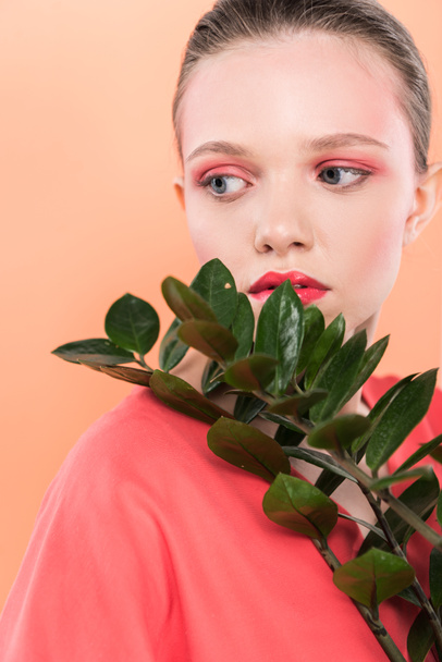 beautiful stylish girl looking away and posing with plant isolated on living coral - Φωτογραφία, εικόνα