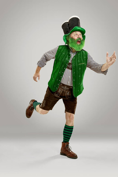 A smiling happy senior man in a leprechaun hat with beard running at studio. He celebrates St. Patricks Day. The celebration, festive, beer, holiday, alcohol, party concept - Photo, Image