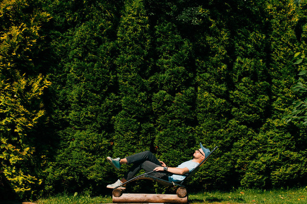 man relaxing in chaise longue beyond swimming pool at villa in Sicily with wall of green southern trees on background - Фото, зображення