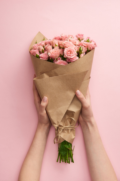 cropped shot of female hands holding bouquet of beautiful pink roses wrapped in craft paper on pink - Фото, зображення