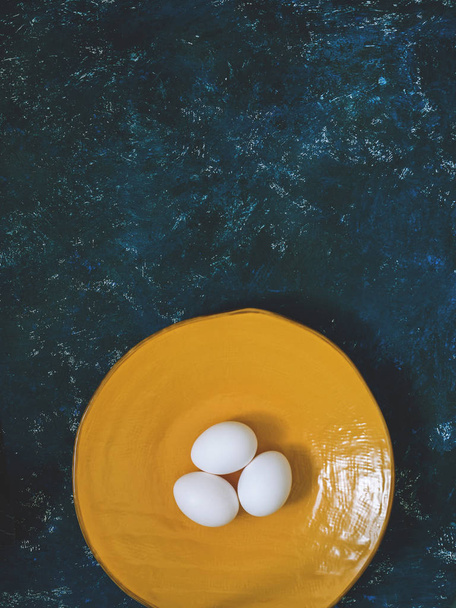 Boiled chicken eggs on a yellow plate. The concept of fertilization, resurrection, Easter. Blue background. Flat lay, top view - 写真・画像