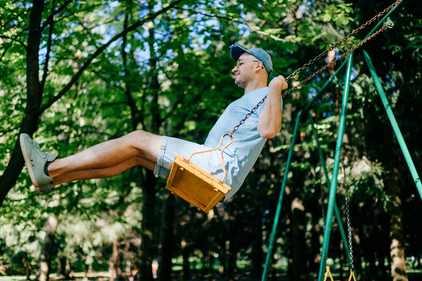 adult man swinging on swing on playground in summer park - Foto, Imagem