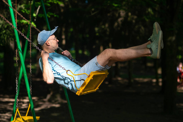 adult man swinging on swing on playground in summer park - Foto, afbeelding