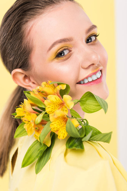 sonriente chica elegante con flores mirando a la cámara con centro de atención en el fondo
 - Foto, Imagen