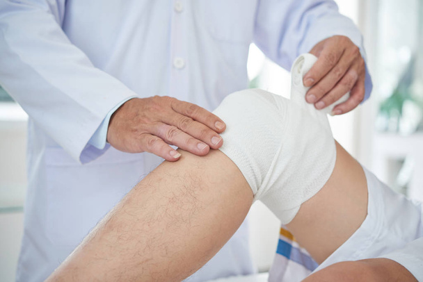 Unrecognizable medical practitioner wrapping bandage around knee of crop patient - Fotoğraf, Görsel