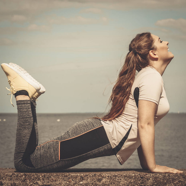 Super fit attractive young woman wearing fashionable outfit working out being active outside during sunny weather. Stretching her back or practice yoga next to sea - Fotoğraf, Görsel