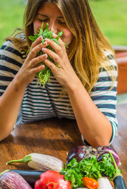 Glimlachende vrouw zitten met hoop verse groenten en ruiken bos van kruiden - Foto, afbeelding