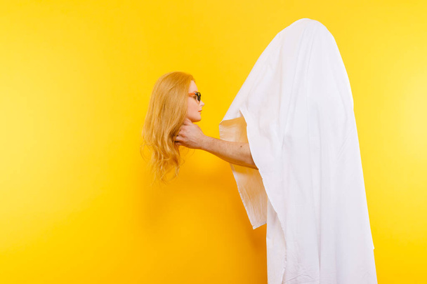 partial view of man covered with white fabric holding plastic mannequin head on yellow backdrop - Foto, imagen